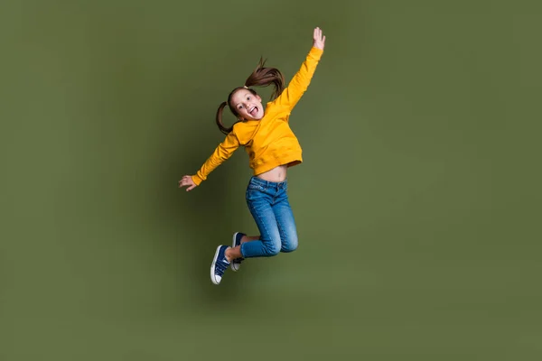 Full length photo of good mood kid with ponytails hairdo dressed yellow sweatshirt denim pants jumping isolated on khaki color background.