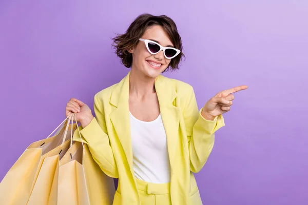 Photo of cute funky lady wear suit blazer dark glasses holding bargains pointing empty space isolated violet color background.
