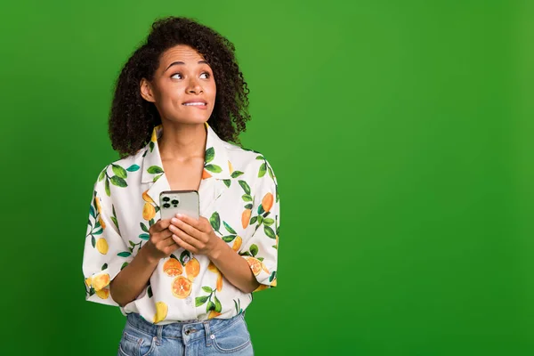 Photo of doubtful unsure lady wear print shirt biting lip typing modern device looking empty space isolated green color background.