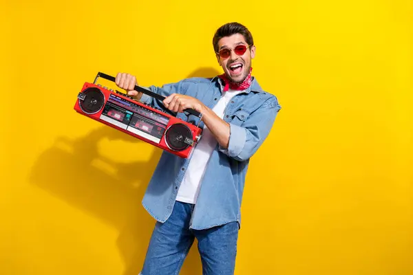 Portrait of positive man with stubble dressed denim shirt sunglass holding boombox at party isolated on vibrant yellow color background.