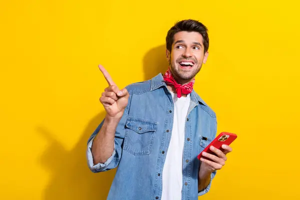 Photo portrait of nice young man hold gadget point look empty space dressed stylish denim garment isolated on yellow color background.