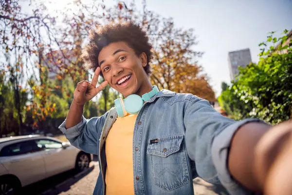 Photo of funky cheerful guy dressed jeans shirt headphones showing v-sign cover eye tacking selfie outdoors urban city park.