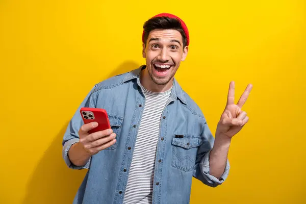 Photo of excited funky guy wear denim jacket showing v-sign chatting modern device isolated yellow color background.