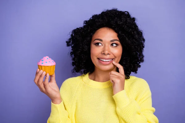 Photo of minded woman with afro hairdo dressed yellow sweater look at tasty cupcake finger on teeth isolated on violet color background.