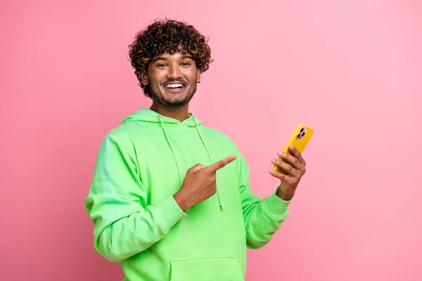 Photo of young student man wearing green sweatshirt pointing finger betting app in smartphone free spins isolated on pink color background.