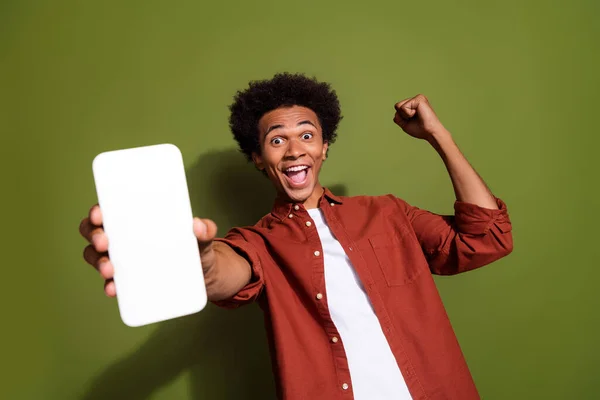 Photo of ecstatic man with afro hair dressed brown shirt showing you smartphone display win betting isolated on khaki color background.