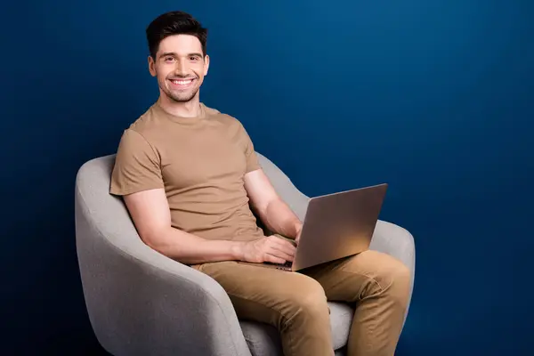 Portrait of smart pleasant man with stubble wear beige outfit sit on armchair with laptop at office isolated on dark blue color background.