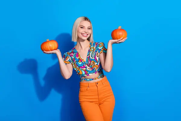 Portrait of toothy beaming girl with straight hairstyle wear stylish blouse holding two pumpkin in arms isolated on blue color background.