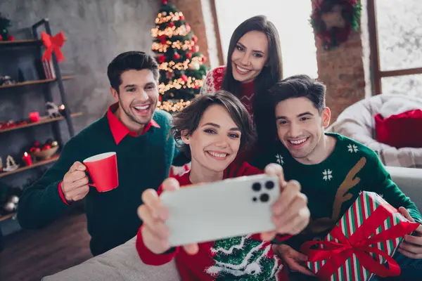 Portrait of group excited funny buddies sitting couch hold smart phone take selfie enjoy hot chocolate receive christmas giftbox indoors.