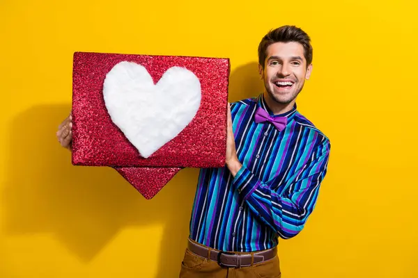 Photo of positive good mood man dressed striped shirt bow tie holding big social media heart box isolated on yellow color background.
