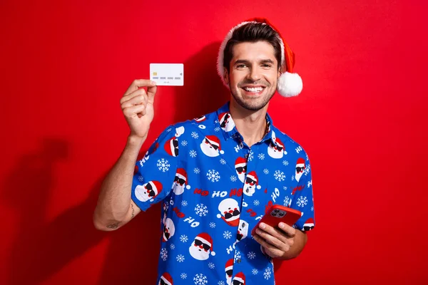 クリスマスの帽子の印刷物のシャツを身に着けている男の写真 スマートフォンは鮮やかな赤い色の背景で隔離されたクレジットカードを示します — ストック写真