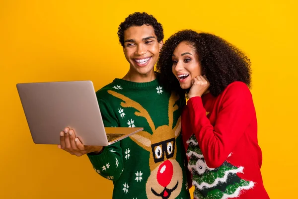 Portrait of two positive funny people hold wireless netbook online shop new year offer isolated on yellow color background.