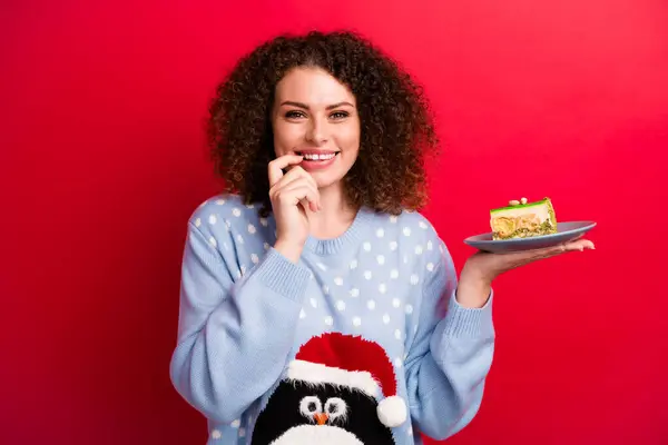 照片上穿着丑陋的圣诞羊毛衫 笑容可亲的小女孩 手指头嘴在煮美味的蛋糕 孤立的红色背景 — 图库照片