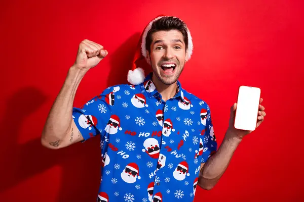 楽しんだファンキーな男の写真は クリスマスの帽子プリントシャツショースマートフォンスクリーンウィン宝くじを鮮やかな赤い背景に分離しました — ストック写真