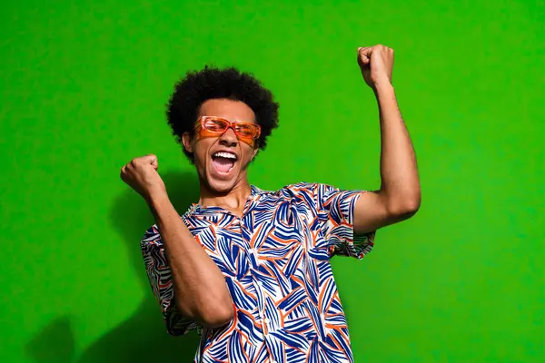 Photo of lucky good mood man dressed print shirt dark glasses rising fists shouting yeah isolated green color background.