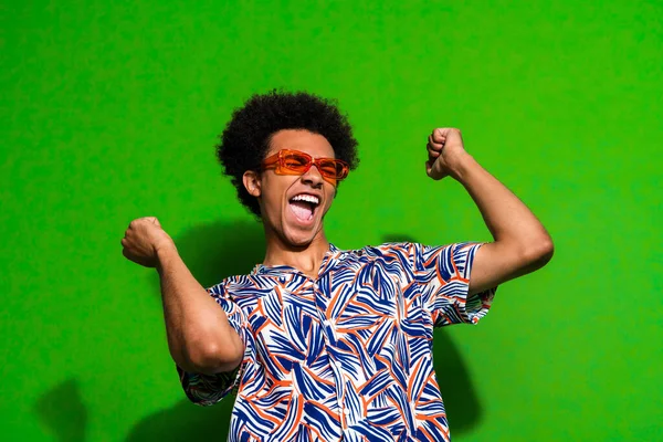 Photo of positive lucky man dressed print shirt dark glasses rising fists dancing isolated green color background.