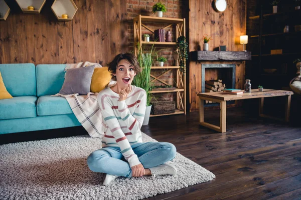 Full size portrait of astonished minded girl sit carpet floor arm touch chin unbelievable reaction wooden interior apartment inside.
