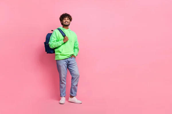 Full body photo of optimistic student wavy haired guy wearing green hoodie with jeans bring rucksack isolated on pink color background.