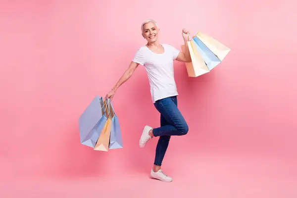 満足した女性のフルサイズの写真は ショッピングバッグを保持している白いTシャツジーンズを着用し ピンク色の背景に隔離された大きな販売を祝います — ストック写真