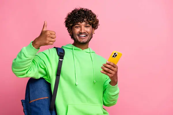 Portrait of optimistic new iphone user young arabian curly haired guy shows like symbol thumb up isolated on pink color background.