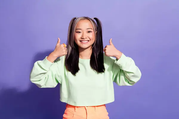 満足のいくアジアの女の子の肖像画 ポニーテールを着用オーバーサイズプルオーバー バイオレット色の背景に隔離された親指を示す2つの腕 — ストック写真