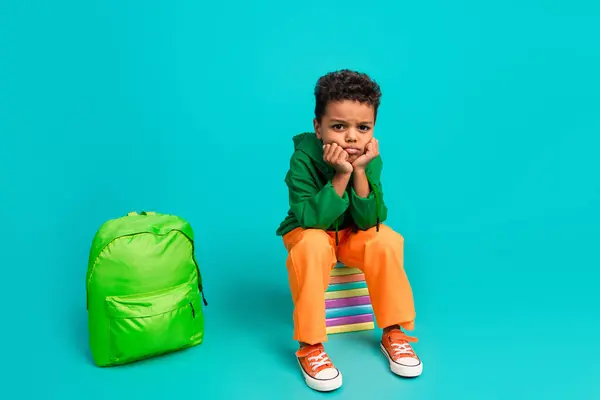 Full size photo of boring tired little boy wear orange pants sit on book near backpack hands on cheeks isolated on teal color background.