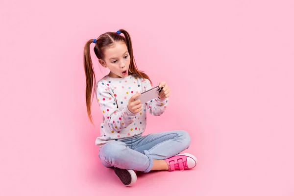 感銘を受けた女子高生のフルサイズの写真は ピンク色の背景に隔離されたスマートフォンのビデオゲームでプルオーバーシットプレイします — ストック写真