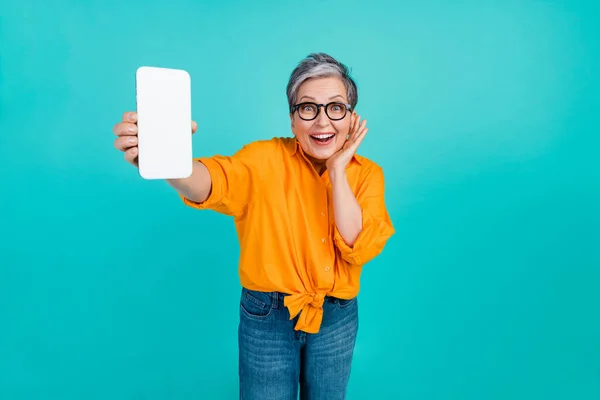Photo of amazed funny business woman in orange shirt holding cellphone display ready for eshopping isolated on blue color background.