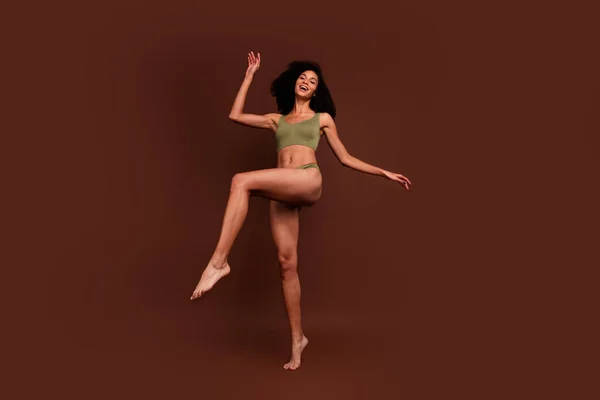 No filter full length photo of cheerful positive sporty girl with thin slim body raise leg isolated on brown color background.