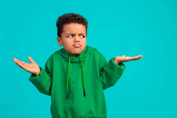 Photo of confused funny schoolboy with wavy hair dressed green pullover look empty space shrug shoulders isolated on teal color background.