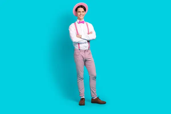 Full length photo of optimistic man dressed pink retro cap pants on suspenders hold arms crossed isolated on blue color background.