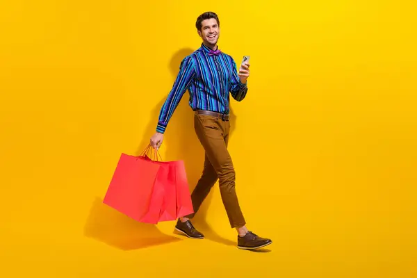 Full body photo of positive man wear bow tie stylish shirt hold shopping bags walk with smartphone isolated on yellow color background.