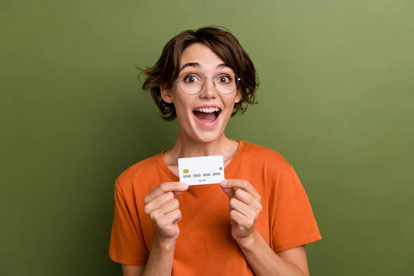 Photo of ecstatic overjoyed girl with bob hair dressed orange t-shirt in glasses presenting debit card isolated on khaki color background.