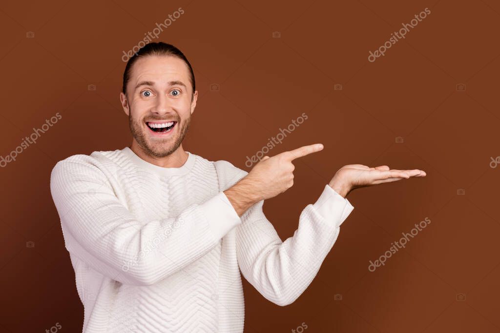 Photo of positive impressed man wear white sweater holding arm showing finger empty space isolated brown color background.