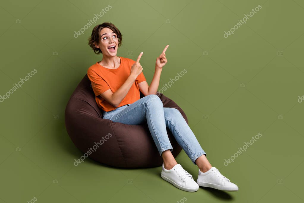 Full length photo of impressed girl dressed t-shirt in eyewear sit on pouf directing look empty space isolated on green color background.