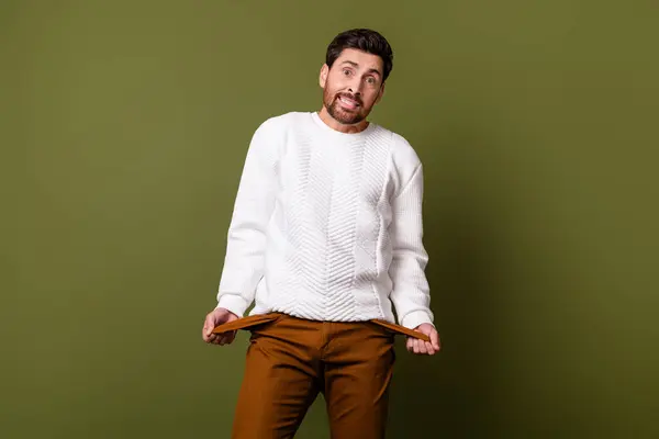 Photo of confused puzzled man wear trendy clothes showing empty pockets no money isolated on khaki color background.