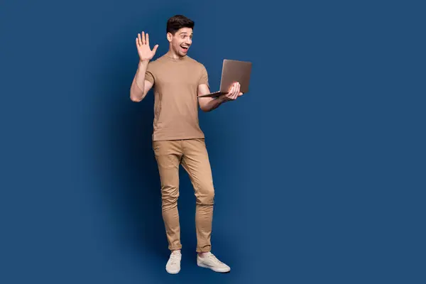 Full length photo of cheerful man wear beige stylish outfit talk on laptop videocall waving hand say hi isolated on dark blue background.