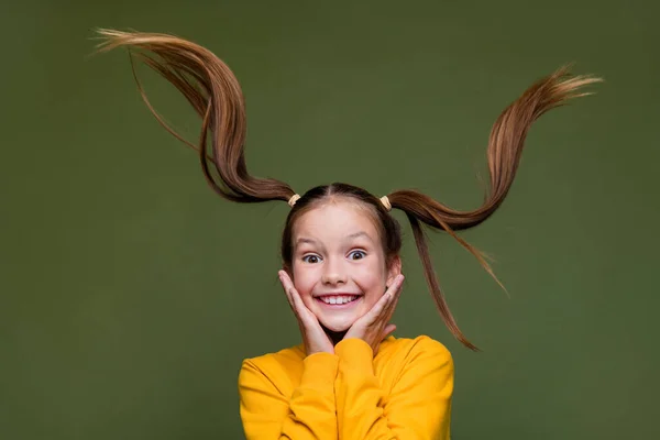 Portrait of optimistic kid with fluttering hair dressed yellow sweatshirt arms on cheeks smiling isolated on khaki color background.