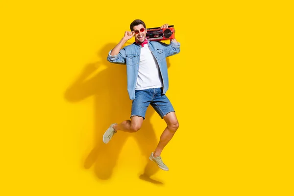 Full body photo of cheerful man dressed denim shirt flying touch glasses hold boombox at summer event isolated on yellow color background.