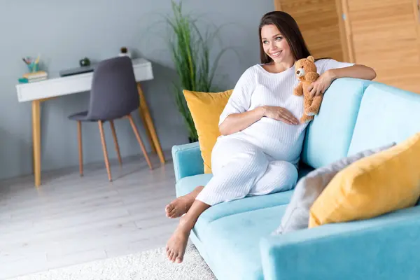 愛らしい夢のような美しい陽気な妊娠中の少女の写真は 新生児と会うために待っているテディベアと快適なソファに座っています — ストック写真