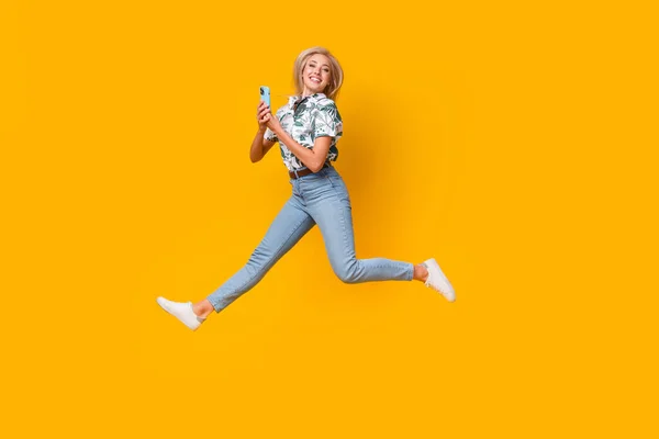 Full length photo of optimistic woman wear blouse denim pants jumping with smartphone in hands isolated on yellow color background.