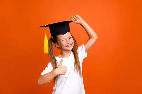 Portrait of clever funny small girl hand touch mortarboard hat demonstrate thumb up isolated on orange color background.