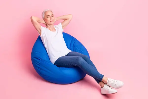Full body photo of satisfied person dressed stylish t-shirt denim pants laying on pouf arms behind head isolated on pink color background.
