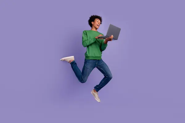 Full length photo of cool excited guy wear green pullover working modern gadget jumping empty space isolated purple color background.