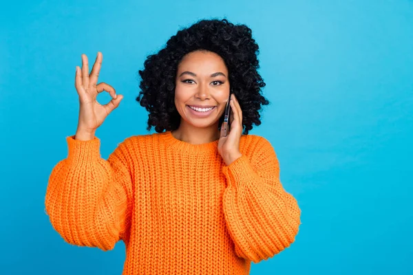 Photo portrait of young woman talk phone show okey symbol dressed stylish knitted orange clothes isolated on blue color background.