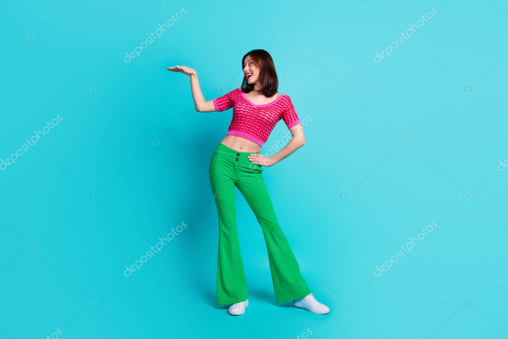 Full size photo of lovely ecstatic girl wear pink top green trousers look at object on arm empty space isolated on blue color background.