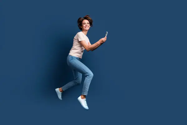 Full length photo of lovely young lady running hold telephone hurry shopping dressed white garment isolated on dark blue color background.