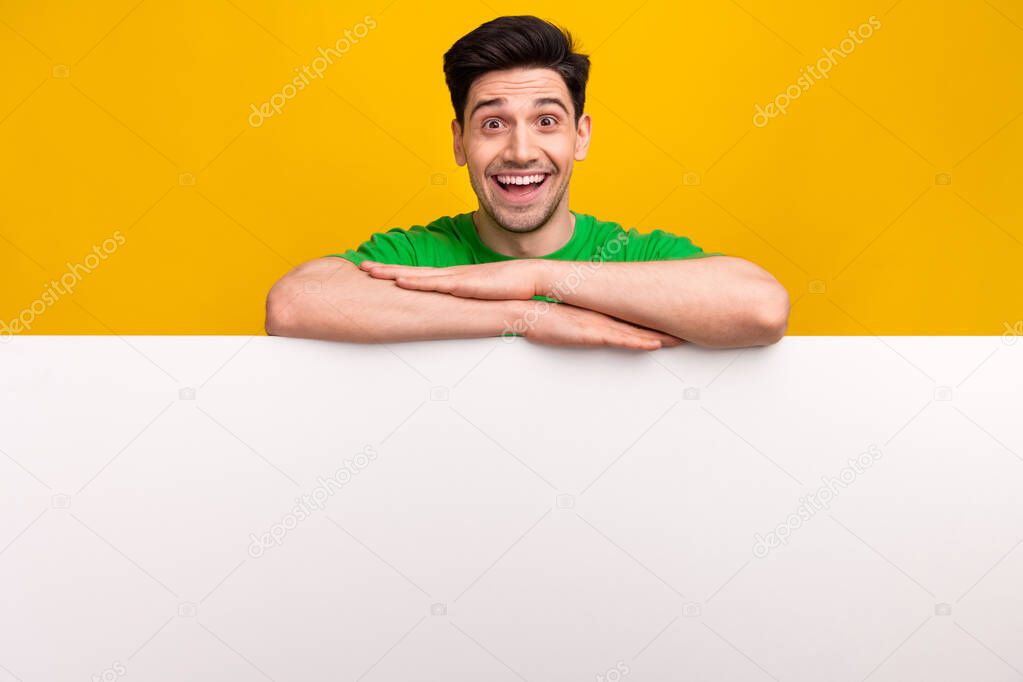 Portrait of astonished overjoyed guy wear stylish t-shirt hold arms on white board empty space isolated on yellow color background.