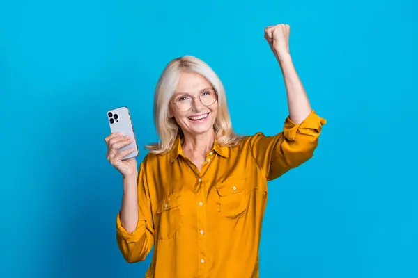 stock image Photo of pretty retired female gadget raise fist celebrate achievement wear trendy yellow silky outfit isolated on blue color background.