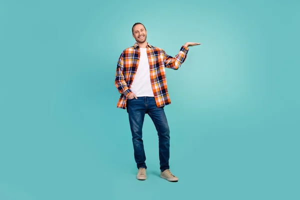 Full length photo of positive man wear flannel shirt jeans arm demonstrate offer empty space isolated on turquoise color background.